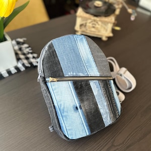 May include: A denim backpack with a vertical striped design in shades of blue and black. The backpack has a zippered closure with a gold-colored zipper and a small handle. A small white accessory is attached to the side.