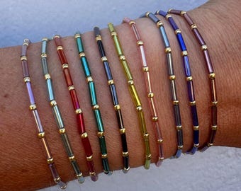 May include: A collection of colorful beaded bracelets. Each bracelet features small, rectangular beads in various colors, including red, green, blue, and purple, separated by small gold-colored beads. The bracelets are worn on a wrist.