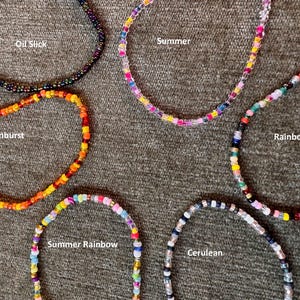 Può includere: Una collezione di collane di perline in vari colori, tra cui "Oil Slick", "Summer", "Sunburst", "Rainbow", "Summer Rainbow" e "Cerulean". Ogni collana è realizzata con piccole perline colorate, con i nomi dei colori scritti sull'immagine.