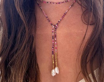 May include: A layered necklace with a beaded choker and a longer strand in a Y-shape. The necklace combines pink, purple, and gold beads, finishing with two white pearl drops. The jewellery is worn on a person.