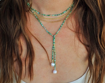 May include: A multi-strand necklace with green, blue, and gold beads. The necklace includes a lariat design with two white pearls. The necklace is worn over a white top. The beads are small and delicate.