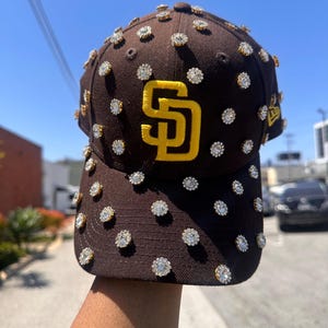 Può includere: Un cappello da baseball marrone con un logo "SD" dorato e strass.