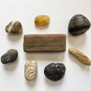 May include: An assortment of smooth, natural stones in various shapes and colors. The collection includes a rectangular, reddish-brown stone, a dark, rounded stone, and several smaller stones with speckled and striped patterns. The stones are arranged on a white surface.