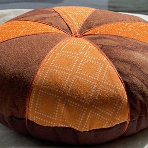 May include: A round, handmade cushion with a pumpkin-like design. The cushion features alternating sections of brown and orange fabric, with a patterned orange fabric in the center. The cushion is approximately 12 inches in diameter.