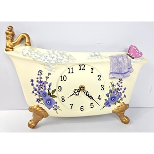 Könnte beinhalten: Eine cremefarbene, badewannenförmige Uhr mit goldenen Klauenfüßen und einem goldenen Wasserhahn. Das Zifferblatt zeigt schwarze Ziffern, während das Äußere mit violetten Blumenmustern, einem geformten violetten Handtuch und einem rosa Schmetterling verziert ist.