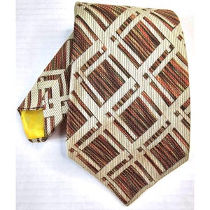 Puede incluir: Una corbata vintage con un patrón geométrico en tonos marrón, naranja y crema. La corbata presenta un diseño repetitivo de líneas y rectángulos que se cruzan. La punta de la corbata tiene un forro amarillo.