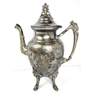 Puede incluir: Una cafetera de metal plateado con una tapa ornamentada con un remate, un pico curvo y un asa con detalles decorativos. El cuerpo está decorado con patrones florales en relieve y se apoya sobre tres patas esculpidas con diseños complejos.