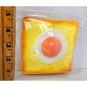 May include: A square slice of yellow toast topped with a fried egg featuring a bright orange yolk, encased in a clear plastic bag. A wooden ruler placed vertically to the left shows the toast measures approximately 4 inches in height.