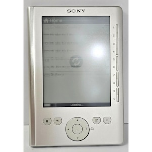 Puede incluir: Un lector electrónico Sony de color plateado con una pantalla rectangular que muestra un menú de inicio con texto que incluye Home, Books by Title, Books by Author y Settings. El dispositivo tiene una columna vertical de botones numerados del 0 al 9.