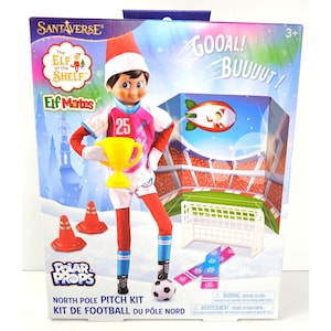 May include: A Santaverse Elf on the Shelf North Pole Pitch Kit featuring a doll in a pink and white soccer uniform with the number 25, holding a yellow trophy. The box includes a soccer ball, two orange cones, a white goal, and a scarf.