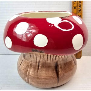 May include: A ceramic mushroom-shaped planter with a red top and white polka dots. The base is brown with vertical textured lines. The planter has a wide opening and is designed for decorative use.