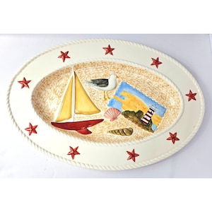 May include: An oval ceramic serving platter with a cream-colored rim featuring a rope-like texture and red starfish accents. The center displays a painted scene with a yellow sailboat, a seagull, seashells, and a red and white lighthouse.