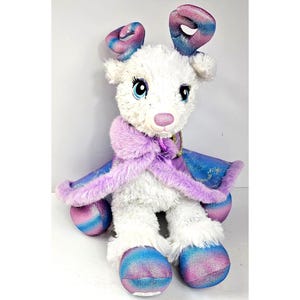 May include: A plush reindeer toy with white fur, large blue eyes, and a pink nose. It features iridescent blue and pink antlers and hooves. The reindeer wears a purple cape with a faux fur collar and gold star patterns on the fabric.