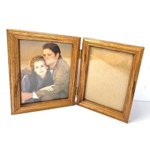 Op de afbeelding: Een scharnierende dubbele fotolijst van lichtbruin hout met een zichtbare nerf. De linkerkant bevat een foto van een persoon met lang haar die een andere persoon in een beige jas omhelst. De rechterkant is leeg met een beige achtergrond.