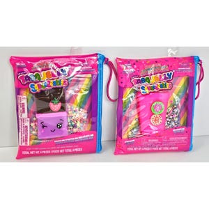Op de afbeelding: Twee Taba Jelly Squeezables sets in roze herbruikbare zakjes. De linker set bevat een bruin en paars chocoladereep-speeltje met een aardbei-icoon. De rechter set bevat een roze bekerspeeltje met ronde decoraties en stervormige strooisels.
