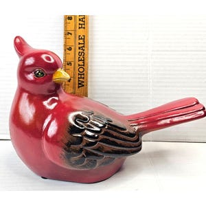 May include: A ceramic cardinal figurine in glossy red with black and brown wing details. The bird has a yellow beak and green eyes. A wooden ruler is placed next to the bird.
