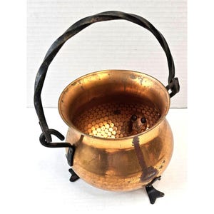 May include: A copper cauldron with a twisted black metal handle and three small feet. The interior of the cauldron is filled with small, round, light-colored objects. The cauldron has a hammered texture and a rustic appearance.