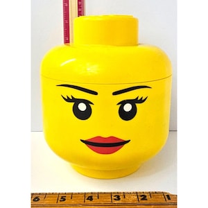 Puede incluir: Un gran contenedor de almacenamiento amarillo con forma de cabeza de Lego. La cabeza tiene cejas negras, pestañas y ojos blancos. Una boca roja cubierta de lápiz labial está pintada en la cara. El contenedor mide aproximadamente 15 cm de alto.