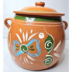 Può includere: Un vaso di terracotta con coperchio, decorato con disegni dipinti in verde, blu e bianco. Il vaso ha un corpo arrotondato e due manici.