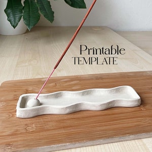 Op de afbeelding: Een witte keramische wierookhouder met een golvende rand, die op een houten oppervlak staat. Een bruine wierookstok brandt in de houder. De tekst "Printable TEMPLATE" is zichtbaar in de afbeelding.