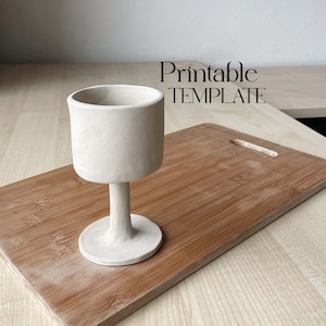 May include: A white ceramic goblet with a wide base and a tall, cylindrical bowl. The goblet is sitting on a wooden cutting board. The text "Printable TEMPLATE" is visible in the background.