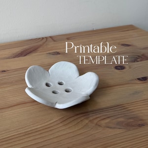 May include: A white, flower-shaped soap dish with three drainage holes. The dish is made of a matte material and sits on a wooden surface. The text "Printable TEMPLATE" is in the background.