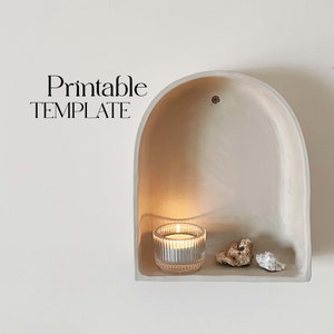 May include: Printable template for a wall niche with a rounded top. The niche is made of white plaster and has a small shelf. A candle is lit inside the niche, and there are two small white stones on the shelf.