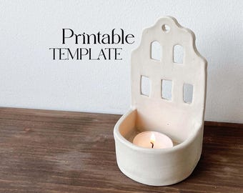 May include: White ceramic candle holder shaped like a house with cut-out windows. A small tea light candle is burning inside the holder. The text "Printable TEMPLATE" is visible in the image.