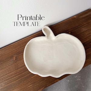 以下が含まれることがあります： 白いセラミック製の、かぼちゃの形をしたお皿。お皿は木製の表面の上に置かれています。お皿の上に「Printable TEMPLATE」と書いてあります。