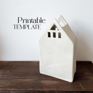 May include: Printable template for a white ceramic house-shaped vase with cut-out windows. The vase is sitting on a dark brown wooden surface.