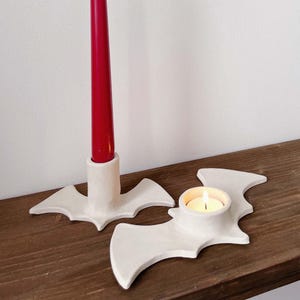 May include: White ceramic candle holders shaped like bats. One holder has a red candle and the other has a small white candle.