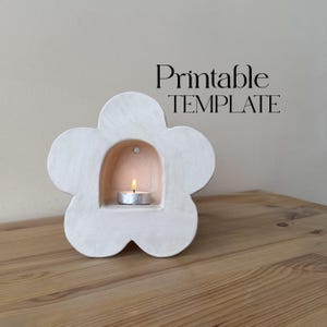 Puede incluir: Un portavelas blanco en forma de flor con una pequeña vela de té encendida en su interior. El portavelas tiene un interior redondeado y está sobre una superficie de madera. El texto "Printable TEMPLATE" se muestra en negro.
