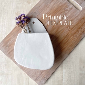 May include: A white ceramic wall planter with a rounded shape and a hole for hanging. The planter is on a wooden chopping board with a text overlay that reads "Printable TEMPLATE".