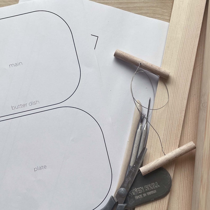 May include: A white paper template with three oval shapes labeled "main", "butter dish", and "plate". The template is on a wooden surface with tools for leather crafting, including a wooden dowel, a leather awl, and a pair of scissors.