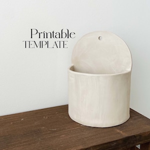Puede incluir: Un macetero de pared de cerámica blanca con la parte superior redondeada y un agujero para colgar. El macetero tiene una superficie lisa y ligeramente texturizada. El texto Printable TEMPLATE es visible en la imagen.