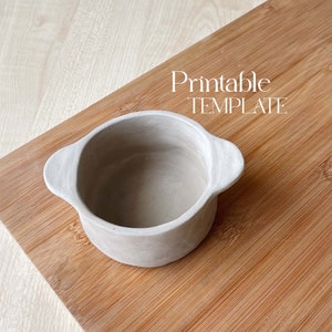 Puede incluir: Un macetero de cerámica gris con dos asas. El macetero está sobre una superficie de madera. Printable TEMPLATE está escrito en blanco sobre la superficie.
