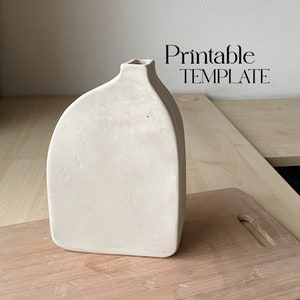 Puede incluir: Un jarrón de cerámica blanco con una forma redondeada y orgánica. El jarrón tiene una abertura ancha en la parte superior y una base estrecha. Está sentado sobre una superficie de madera.