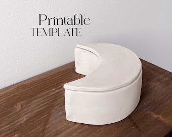 May include: A white ceramic crescent moon shaped box with a lid. The box is sitting on a wooden surface. Printable TEMPLATE is written above the box.