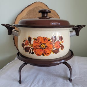 May include: A vintage enamelware pot with a floral design in shades of orange, brown, and white. The pot has a brown lid and sits on a metal stand with three legs.