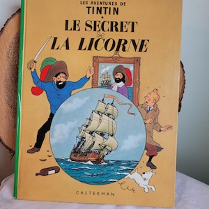 May include: A yellow book cover with the title "Le Secret de La Licorne" in black text. The book is a Tintin adventure by Herg&eacute;. The cover features an illustration of a ship sailing on the sea, a man with a sword, and a dog.