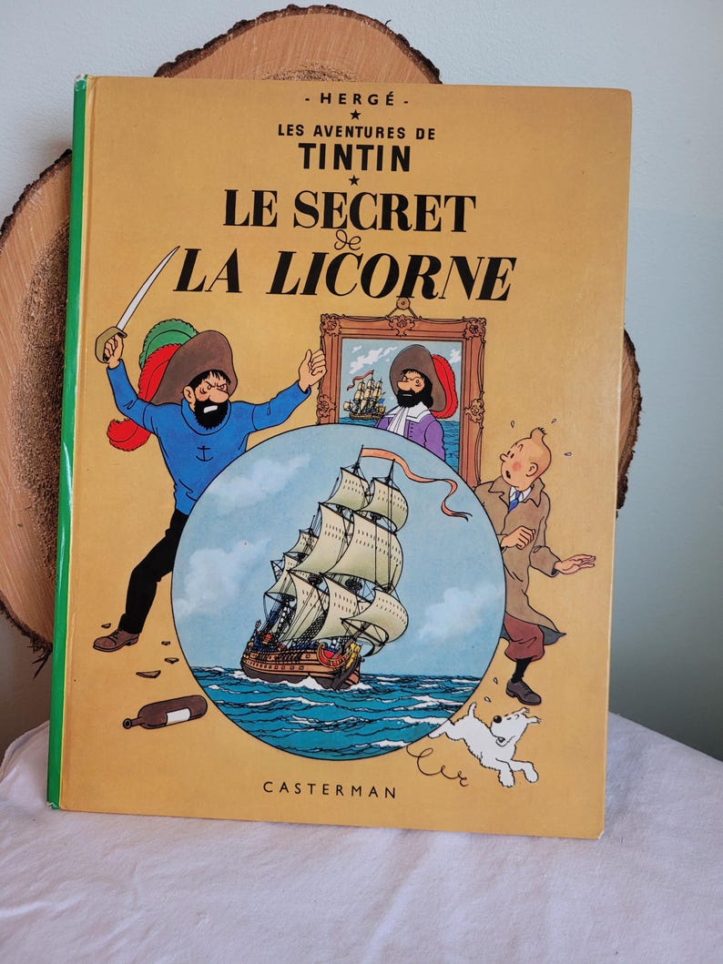 May include: A yellow book cover with the title "Le Secret de La Licorne" in black text. The book is a Tintin adventure by Herg&eacute;. The cover features an illustration of a ship sailing on the sea, a man with a sword, and a dog.