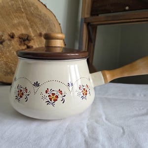May include: Cream-coloured enamel saucepan with a wooden handle and lid. The pan has a floral design in red, orange, and black. The handle and lid knob are made of light-coloured wood. A decorative kitchenware item.
