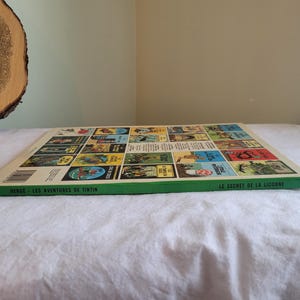 May include: The back cover of a Tintin comic book, "Le Secret de la Licorne" (The Secret of the Unicorn). The cover features a green background with a grid of colorful comic book covers, each depicting a different Tintin adventure. The text "Herge - Les Aventures de Tintin" is printed at the bottom.