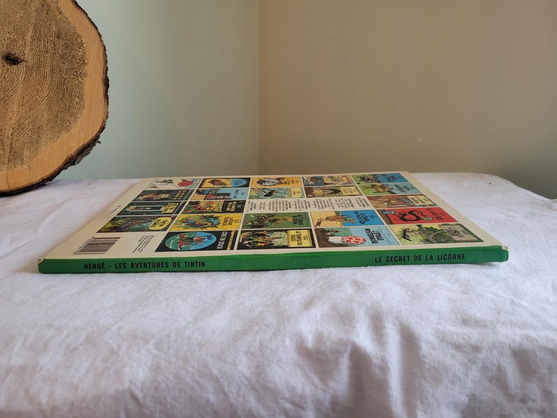 May include: The back cover of a Tintin comic book, "Le Secret de la Licorne" (The Secret of the Unicorn). The cover features a green background with a grid of colorful comic book covers, each depicting a different Tintin adventure. The text "Herge - Les Aventures de Tintin" is printed at the bottom.