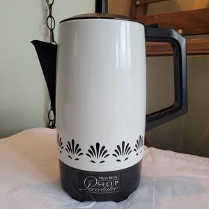 May include: A vintage West Bend 5.9-cup percolator. The coffee maker has a white body with a black base and handle. A decorative black floral pattern adorns the bottom of the white section. The percolator has a black lid and spout.