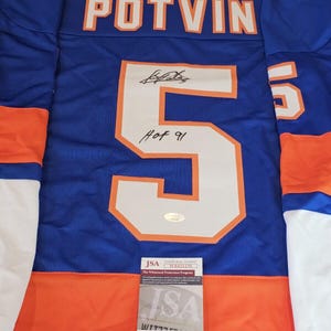 May include: A blue, orange, and white hockey jersey featuring the name POTVIN and the number 5 on the back. The number 5 includes a black ink autograph and the inscription HOF 91. A JSA authentication card is placed over the lower portion.