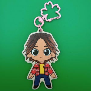 May include: A wooden keychain featuring a cartoon character with brown hair, wearing a red and white patterned shirt, yellow shirt, blue jeans, and a pink flower keychain.