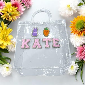 Op de afbeelding: Transparante plastic tas met een roze handvat en roze glitterletters die "KATE" spellen. De tas heeft een paarse konijn en een oranje wortel patch aan de voorkant.