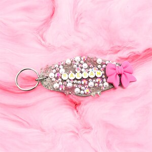 Può includere: Un portachiavi personalizzato argentato con il nome "RAEGAN" in lettere dorate. È decorato con strass e perle. Un fiocco rosa è attaccato. Lo sfondo è rosa marmorizzato. Perfetto per un regalo personalizzato.
