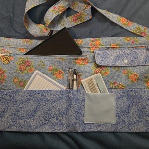May include: A light blue floral print apron-style bag with multiple pockets and a long strap. The bag features a zipper pocket, a small flap pocket, and several open pockets containing a notepad, pens, and a card. The floral pattern includes orange and pink roses.